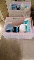 Photo shows two clear plastic bins containing medical supplies including white absorbent roll, unopened packs of sterile dressings, and Omron blood pressure monitor cuff in one bin.