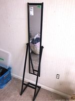 Full view of the standing mirror with its metal frame and adjustable support, showing the mirror in an upright position on the floor against the wall.