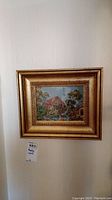Framed needlework hanging on a wall, showing a landscape with a cottage, trees, and a bridge. Frame is gold-colored with ornamental design.