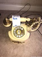 Front view of an off-white rotary dial phone with brass accents, showing handset and coiled cord