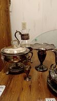 Lot showing silver plate chafing stand with glass lid, glass pitcher with silver plate stand, and silver plate compote on wooden table