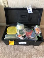 Photo of open tool box showing assortment of various tape rolls inside including clear packing tape, masking tape, duct tape, and double-sided tape. The box is black plastic with a yellow latch and a label 'TAPES' on the front.