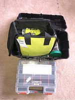 Top view of open black toolbox containing a yellow and black bag holding the Ryobi drill and a clear plastic compartment box labeled with 'Machine Screws'.