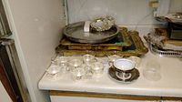 Photo of silver plated tray, wood table tops, metal and glass teacups, metal plates and Bavaria teacup and saucer on white surface.