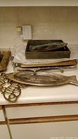 Photo showing a variety of trays including wooden tray with porcelain insert, metal trivet, fish-shaped silverplate tray, and decorative plates stacked on a counter.