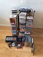 Photo of multiple CDs stacked in wire and wooden holders, cassette tapes in a black case, Sony Walkman with earphones, all placed on a wooden floor.