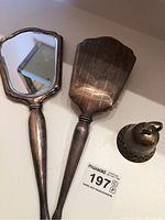 Photo showing the hand mirror, hairbrush, and silver plated bell arranged together on a white surface showing tarnished details and overall set condition.