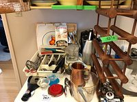 Photo showing a collection of kitchen utensils including cutlery tray with forks, spoons, knives, wooden carving board, electric can opener, wine cooler, ice bucket, and various metal and plastic bowls and utensils.