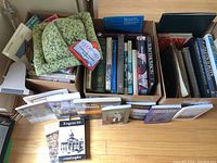 Two cardboard boxes filled with large hardcover and coffee table books, some stacked, visible titles and one cloth bag