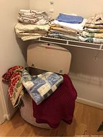 Image showing a retro laundry hamper covered with various blankets including a red knit blanket and a blue patchwork throw, with folded sheets on a wire shelf above
