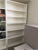 Tall white shelving unit with six shelves and adjustable peg holes, standing empty against a wall next to another shelving unit and cabinet.