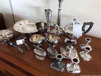Wide view showing silver teapot, cream and sugar serving pieces, candlesticks, small goblet, napkin holders, lighters, and silver dish arranged on wooden surface.
