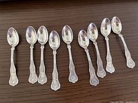 Eight ornate sterling silver coffee spoons arranged horizontally on a wood surface.