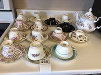 Full lot of 14 vintage teacups and saucers displayed with Royal Albert teapot, showing variety of floral patterns and colors including pinks, yellows, blues, greens, and gold accents.