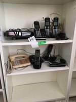 Shelf with various telephones and clock radio, including corded and cordless models, and an Alcatel flip phone.