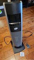 Front view of tall grey and black ceramic tower heater on wood floor with power cord and remote on floor next to base.