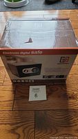 Box of Guardwell electronic digital safe placed on wooden floor with visible slight damage on top of box.