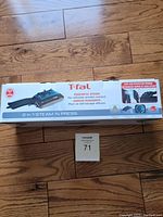 Photo of the original boxed T-fal 2 in 1 Steam 'n Press steamer on wooden floor with lot tag visible.