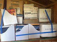 Photo shows a collection of 3 white file boxes secured with blue straps containing books and magazines, stacked with 6-7 additional cardboard boxes behind and on top, some labeled and some plain.