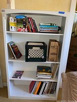 White bookshelf filled with assorted books of various sizes, a black milk crate, and a cardboard box.