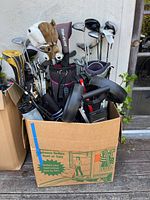 Golf clubs packed in three golf bags within a cardboard box, along with golf caddy parts such as wheels.