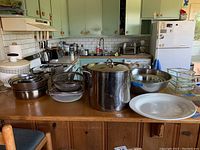 Wide shot showing assortment of kitchenware items including stainless steel crab pot, stacked stainless steel bowls, various Pyrex glass baking dishes, and white ceramic plate on wooden kitchen counter.
