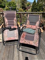 Two reclining lounge chairs on a wooden deck. Chairs have camouflage-style fabric with black mesh sections and wooden armrests. Cushions and a headrest pillow are included.
