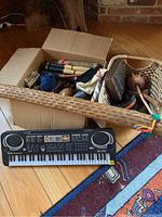 Photo of lot contents showing long wooden rain stick lying on floor, electronic keyboard, cardboard box and woven basket with multiple instruments inside.