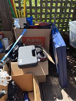 Cardboard boxes and bags filled with sporting gear including a grey hard case, headphones, rollerblades, and protective gear.