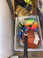 View of tennis racket with basketball and various balls underneath including a rainbow colored ball.
