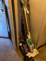 Four vacuum cleaners grouped together in a corner including a green and white upright steam mop vacuum, a brass-colored upright vacuum, and a black handheld vacuum