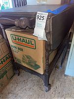 Side view of the tea cart showing turned spindle legs and one side handle, with a U-Haul book box stored beneath the cart. Cart has caster wheels.
