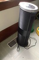 Full view of the black tower air purifier standing on floor near vent, showing overall height and shape.