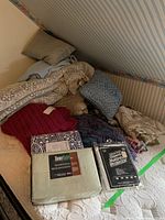 Photo of bed showing sheet set packaging, mattress protector packaging, multiple knitted throws and blankets, and several throw pillows.