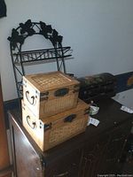 Photo showing two wood storage boxes with metal handles and wicker/rattan paneling stacked on a surface with part of a metal wall shelf visible behind them