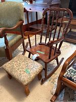 Child-size rocking chair with spindled back next to upholstered footstool on carpeted floor, surrounded by other furniture.