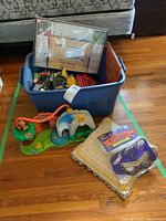 Blue plastic bin filled with assorted children's books and game pieces, framed 'The Game of Quinte' board game, children's toy and pillow with feathered eye mask and ski goggles in front.