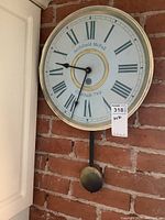 Full view of round vintage wall clock with light blue clock face, black clock hands, Roman numerals, and brass ring detail. The clock is mounted against a brick wall with visible pendulum below.