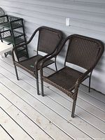 Two brown resin wicker deck chairs placed side by side on a porch with white wooden flooring and light gray siding, showing front and side views.