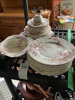 Photo of stacked floral dinner plates, side plates, serving dishes, and a single teacup on a black table with other items in background.
