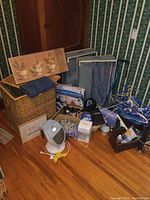 Overview of assorted laundry and household items arranged on floor against wallpapered wall. Includes wicker hamper, framed prints, blue storage bags, boxes, heater, baskets, mop, and hangers.