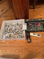 White box filled with approximately 40 collector spoons of various styles and designs on wooden floor.