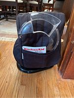 Black stand mixer cover with clear plastic splatter guard attached and KitchenAid instruction book in pocket