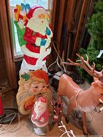 Large Santa blow mold with green sack and blue gloves, standing near a window in wooden interior. Fragile head with tears noted. Other decorations visible around it.