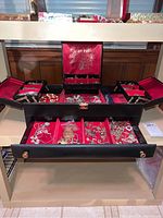 Wide view of open black jewelry box with multiple compartments filled with assorted costume jewelry including bracelets, necklaces, pendants, pins, and earrings on red velvet lining.