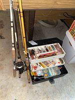 Fishing rods and tackle box on garage floor, showing rods standing and open tackle box with lures visible