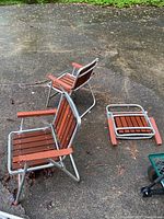 Three folding chairs with metal frames and wooden slats. One chair folded flat, two chairs unfolded. Outdoor setting with some dirt and leaves on chairs and ground.
