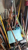 Multiple long-handled garden tools including shovels, rakes, hoes with wooden and colored handles along with a blue plastic snow shovel leaning against the wall