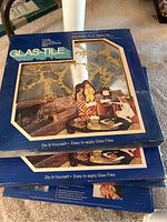 Three stacked blue boxes of Glas-Tile by Hoyne glass tile mirrors, each box containing one dozen 12x12 inch decorative glass mirror tiles with gold crackle design, shown unopened and in original packaging.