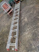 Full view of one metal extension ladder on the ground, silver metal with red end caps and rusted rope securing it.